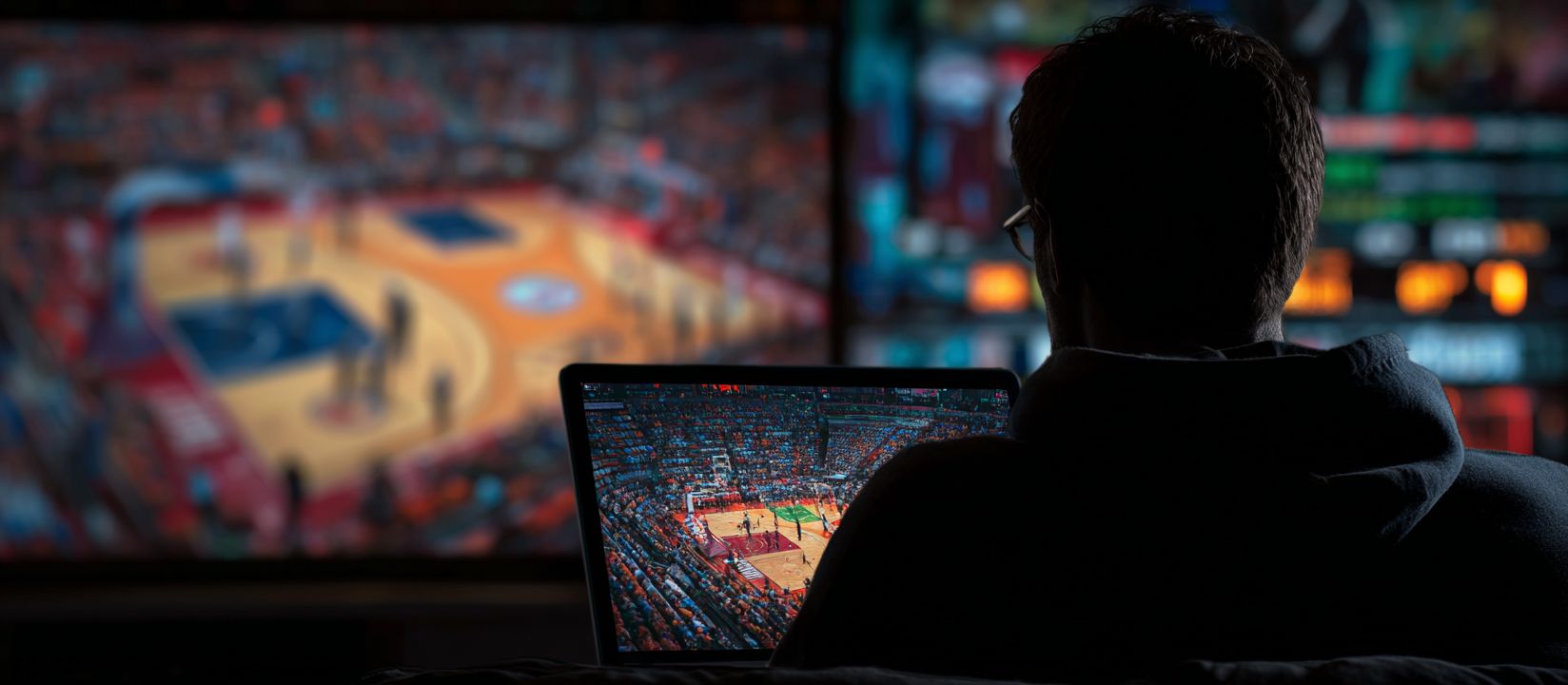 Man watching basketball game live on laptop with big screen in background, representing sports betting at 55WW Casino